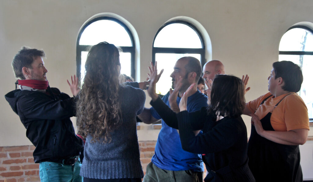 gruppo di otto persone con uno al centro mani aperte all'altezza dei visi e sguardi aperti l'un l'altro